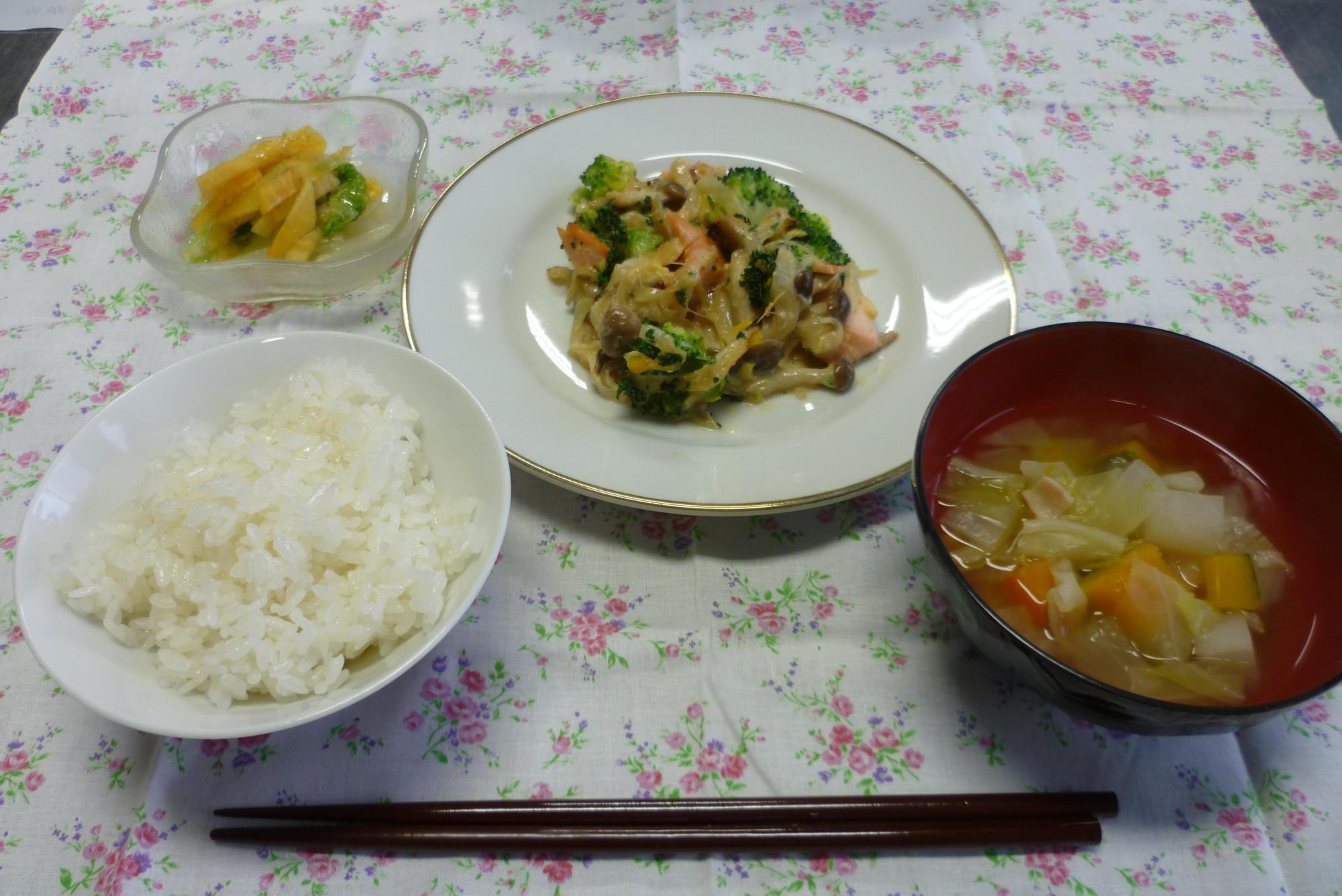 白菜と柿の甘酢和え、鮭のコーンクリーム焼き、野菜がたくさん入ったスープ、白米、漬物がテーブルの上に並べられてある写真
