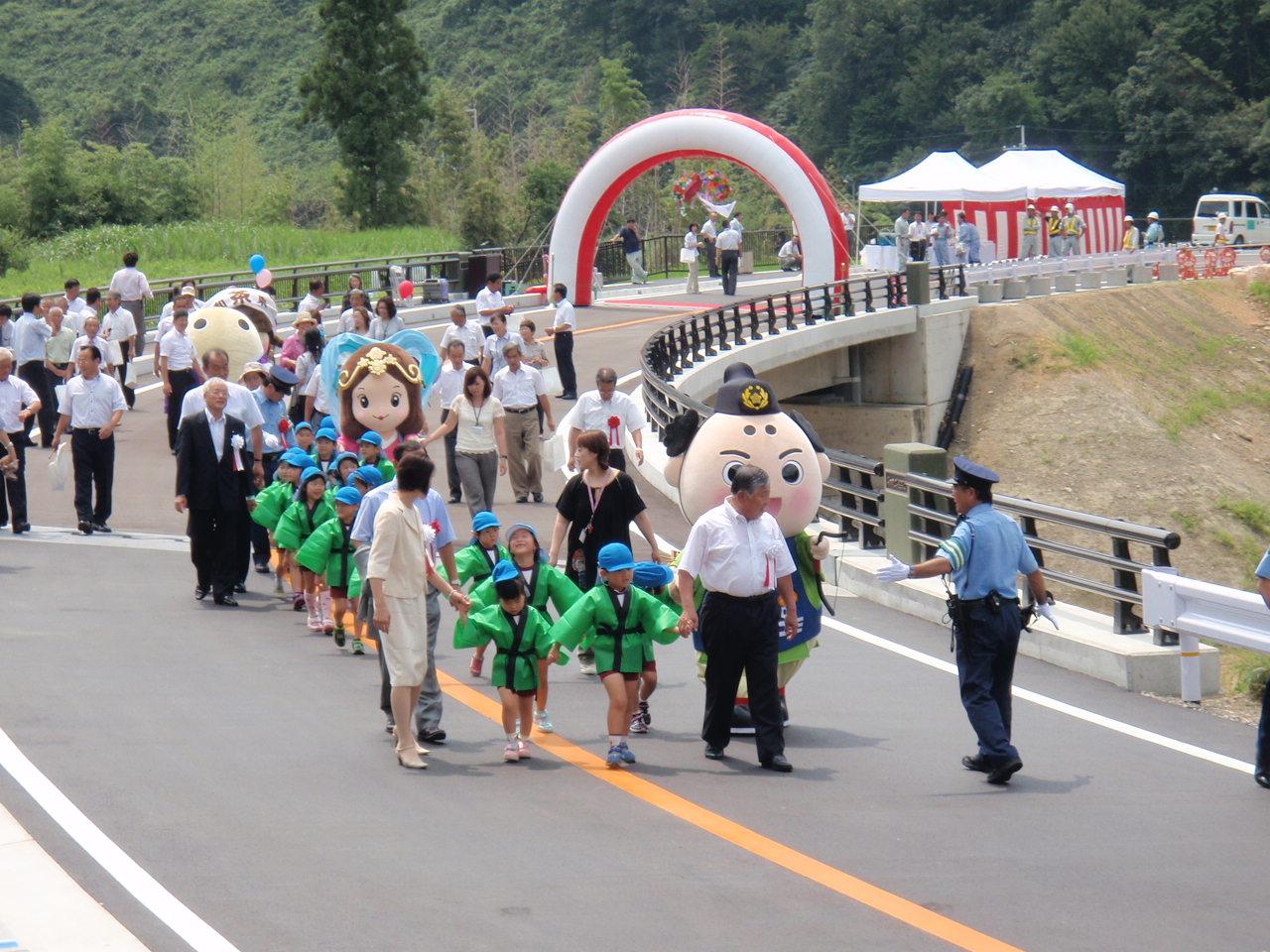 青い帽子に緑色の法被姿の園児たちと和束町長やゆるキャラたちが手を繋いで警察官を先頭に橋を渡っている写真