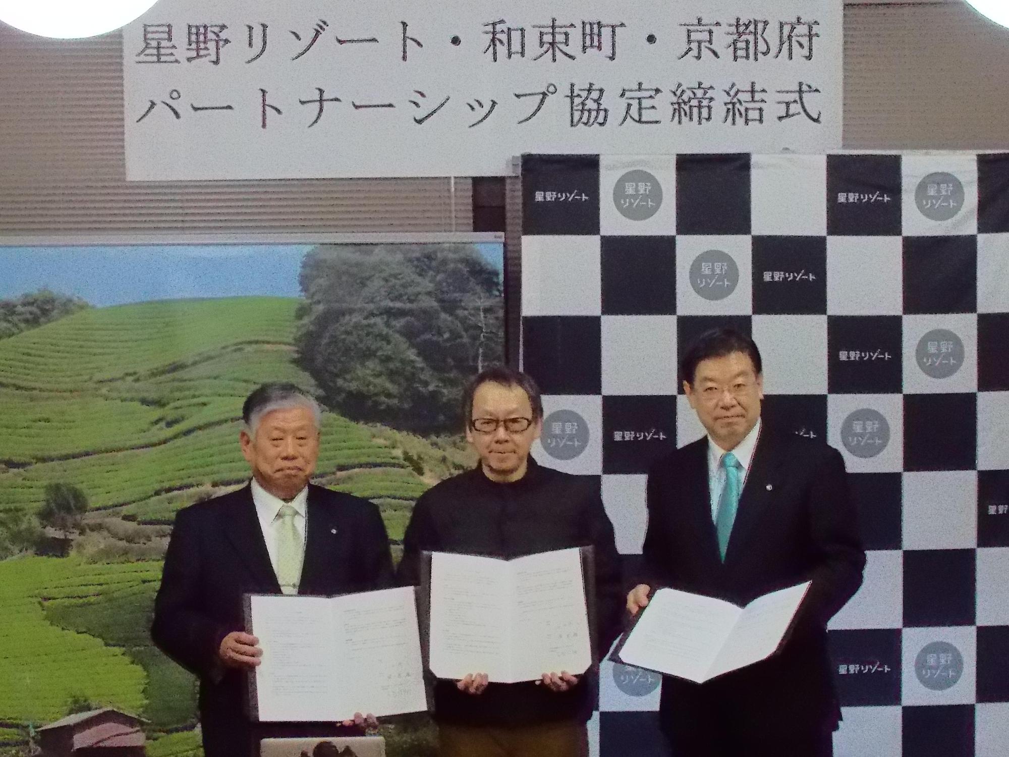 3名の男性関係者たちが協定書を開き、横一列に並んで写っている写真