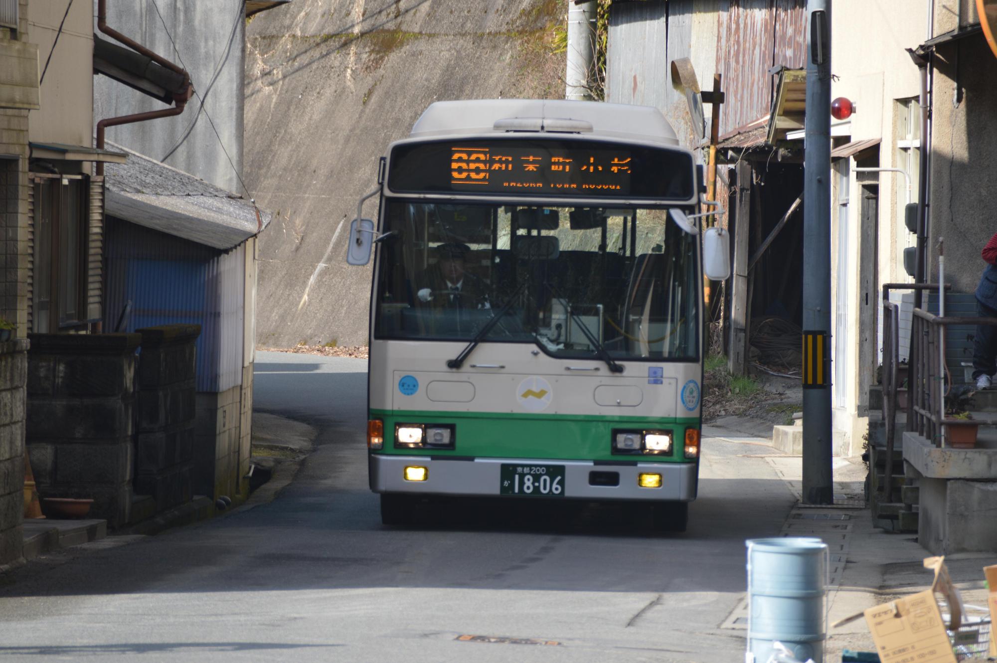 両側に建物が建っており、対向車とすれ違えない道幅の道路を走っているバスの写真