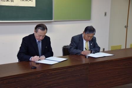 茶色のテーブルの席に座り協定書に署名を行っている町長と菊池理事長の写真