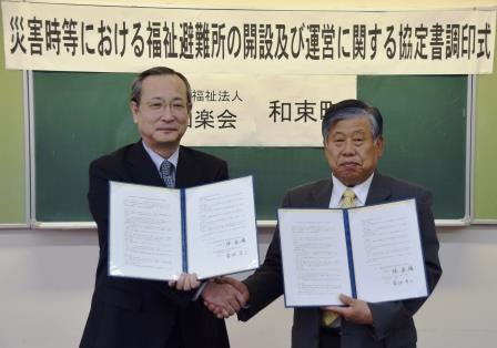菊池理事長と町長がそれぞれ片方の左手で開いた協定書を持ち、もう片方の右手で握手をしている写真