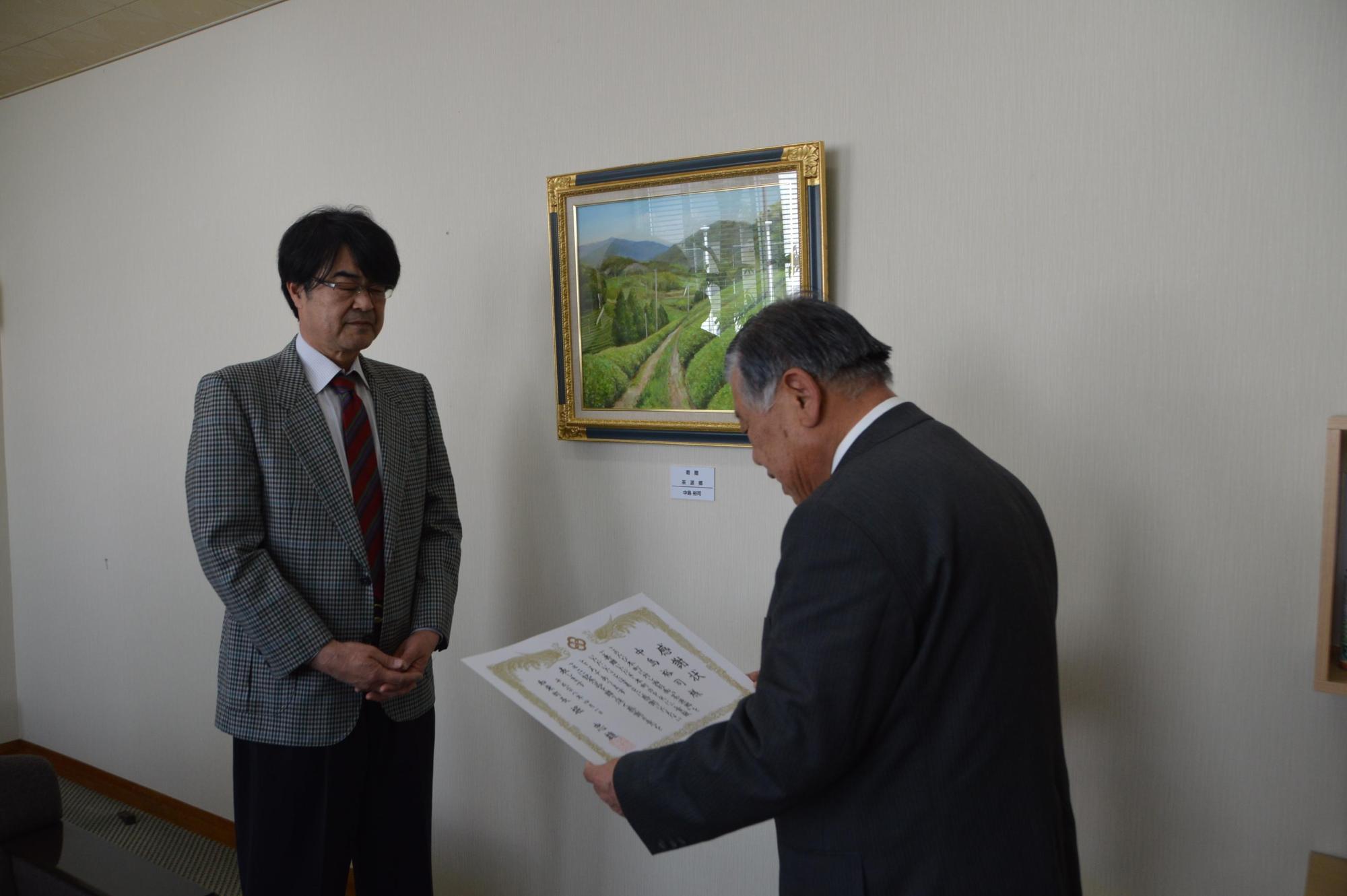 額縁に入った絵が飾られてある壁の前で男性が中島裕司氏に表彰状を読み上げている写真